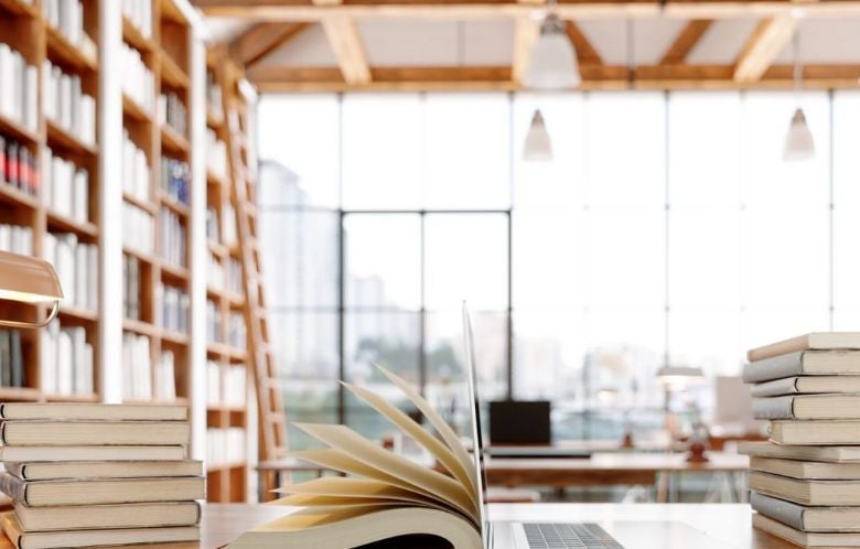 An open library space with many books stacked on a table with a laptop.