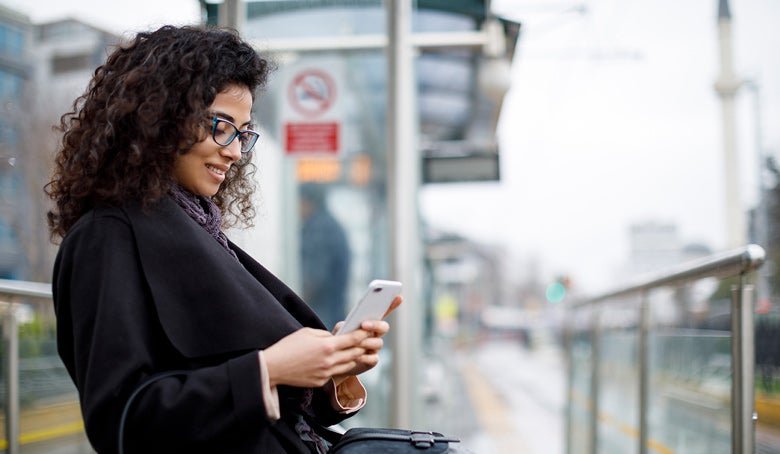 A professional woman reading on her phone while sitting on a bench waiting for her train.